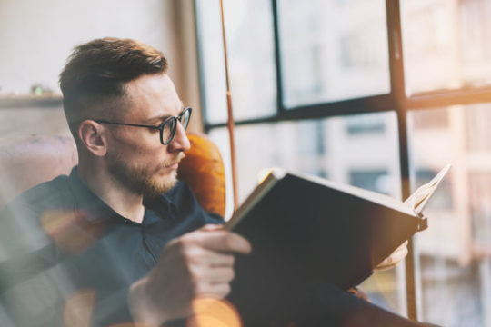 Portrait handsome bearded businessman wearing black shirt.Man sitting in vintage chairmodern loft studio, reading book and relaxing. Blurred background, film effect.